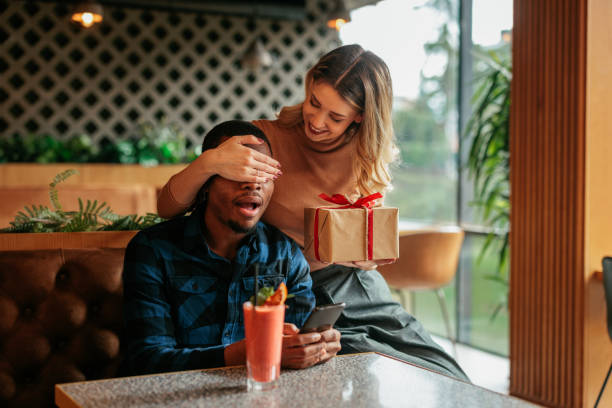 Eine Frau hält ihrem Partner an Valentinstag die Augen zu, während sie ihm eine Geschenkbox überreicht. 💝